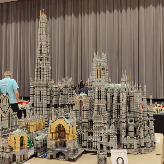 Eine komplizierte Lego-Ausstellung architektonischer Strukturen, einschließlich eines hohen Turms und mehrerer Gebäude, steht auf einem Tisch in einem Raum mit grauen Vorhängen.