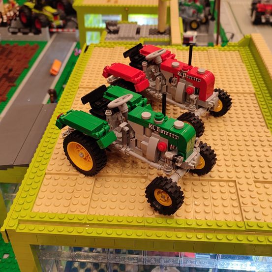 Zwei Spielzeugtraktoren, einer rot und einer grün, stehen auf einer Plattform mit grünem Rand. Sie gehören zu einem Lego-Set, mit anderen Lego-Strukturen im Hintergrund.