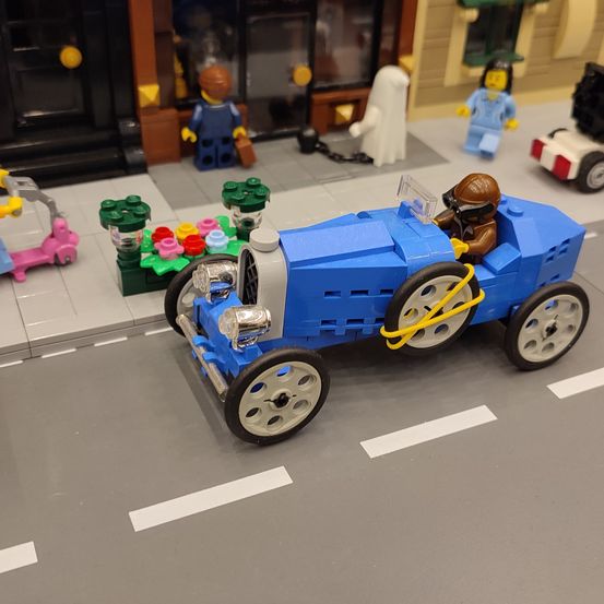 Ein blaues Spielzeugauto mit Fahrer steht auf einer Straße aus Lego. Umgeben ist es von Lego-Figuren, Gebäuden und einem Trolley. Das Auto hat weiße Markierungen auf der Straße.