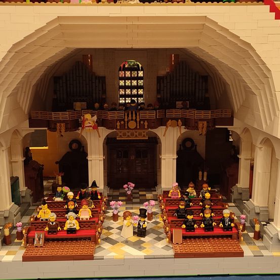 Ein Lego-Modell des Kircheninneren mit Bänken, Altar und einer Orgel. Figuren sitzen auf Bänken, Blumen schmücken den Bereich und ein Buntglasfenster befindet sich darüber.
