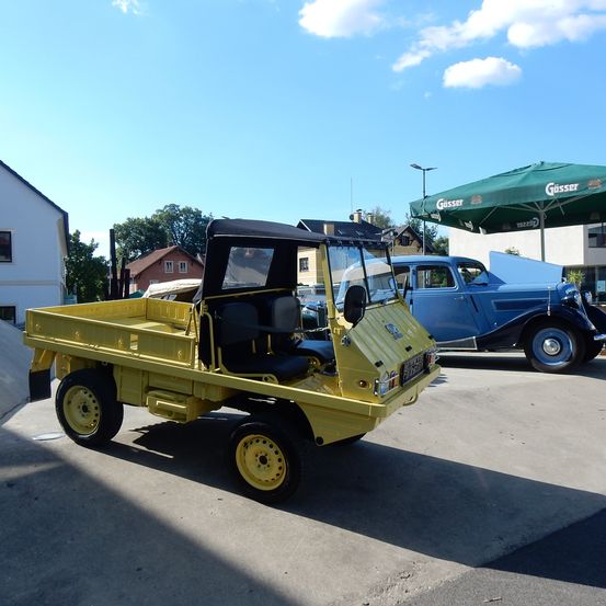 Ein gelbes Oldtimer-Fahrzeug mit schwarzem Dach steht auf einem Parkplatz. Dahinter ist ein blauer Oldtimer. Auf der rechten Seite steht ein grüner Schirm.