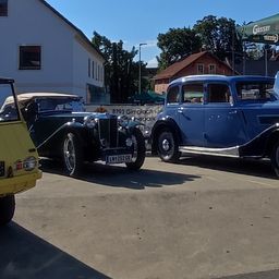 Drei Oldtimer sind auf einem Parkplatz unter einem grünen Schirm geparkt. Ein gelbes Auto links, ein blaues Auto in der Mitte und ein grünes Auto rechts. Ein Fahrrad ist in der Nähe geparkt.