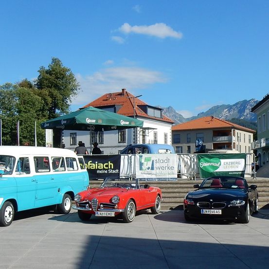 Drei Oldtimer sind vor einem Gebäude mit einem grünen Schirm geparkt. Ein blaues Auto ist links, ein rotes Auto in der Mitte und ein schwarzes Auto rechts. Dahinter stehen zwei Gebäude, eines mit einem roten Dach und das andere mit einem grünen Banner. Im Hintergrund sind Berge zu sehen.
