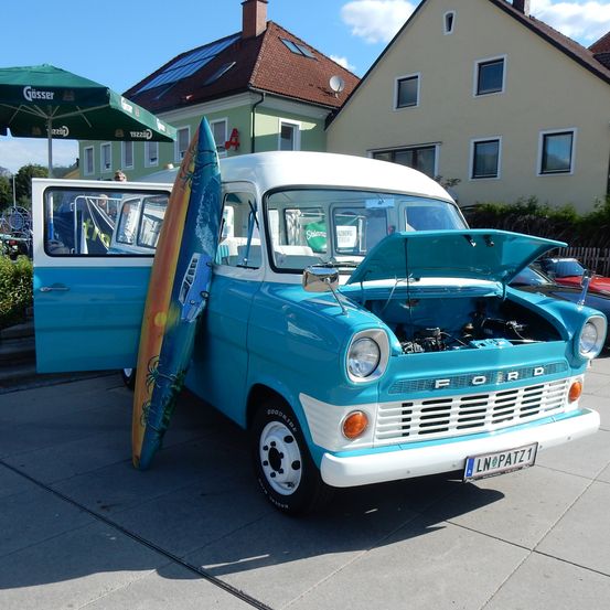 Ein vintage blau-weißer Ford-Van mit offener Tür und einem Surfbrett, das darangelehnt ist, geparkt vor einem Haus mit Solarpanelen.