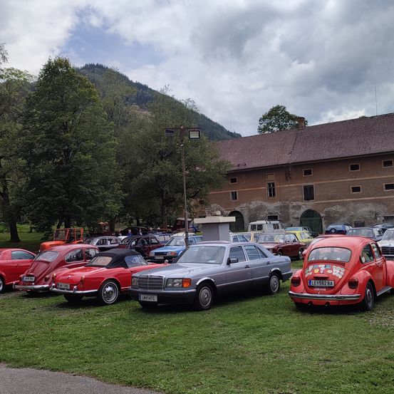 Eine Gruppe von Oldtimern, die auf einem Rasenfeld geparkt sind, mit einem großen Gebäude und Bergen im Hintergrund.