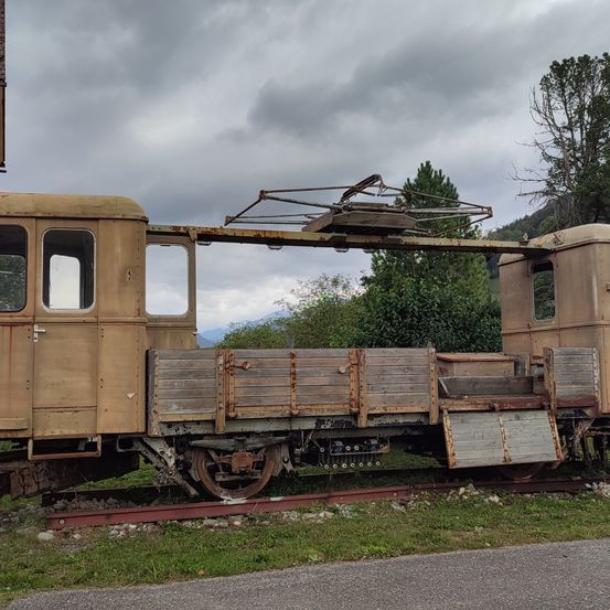 Ein alter, rostiger Eisenbahnwagen steht auf einem Gleis, mit einer Metallstruktur oben drauf. Er ist am Straßenrand geparkt, mit Gras und Bäumen im Hintergrund.