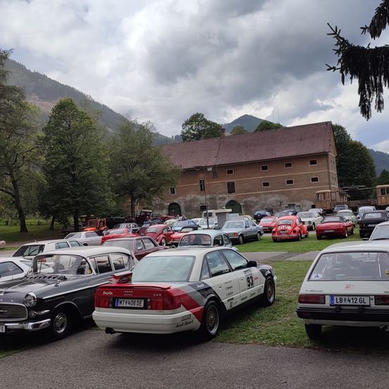 Ein Parkplatz voller Oldtimer, mit einem großen Gebäude und Bergen im Hintergrund.