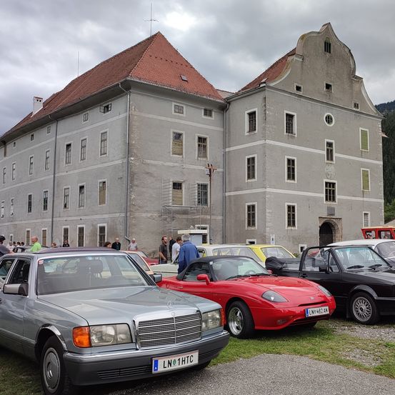 Eine Sammlung von Oldtimern vor einem alten Gebäude mit rotem Dach. Leute stehen herum, einige unterhalten sich.