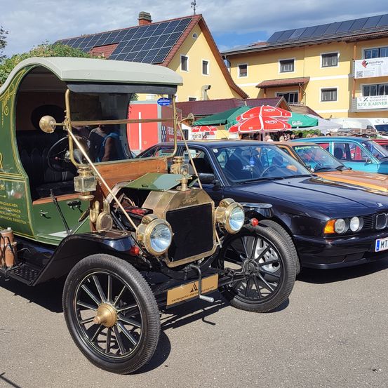 Ein antikes grünes Auto mit goldenen Akzenten ist in einer Reihe von Oldtimern geparkt. Dahinter befinden sich Gebäude mit Solarpanelen und einige Leute unter Schirmen.