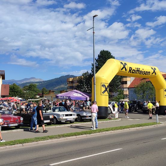 Eine Straßenszene mit einem gelben Bogen, geparkten Autos an der Seite und Menschen, die herumlaufen. Im Hintergrund befinden sich Gebäude und Berge.