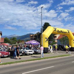 Eine Straßenszene mit einem gelben Bogen, geparkten Autos an der Seite und Menschen, die herumlaufen. Im Hintergrund befinden sich Gebäude und Berge.