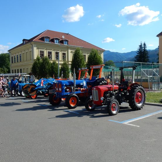 Eine Reihe von Oldtimer-Traktoren, die auf einem Parkplatz mit einem Gebäude und Bergen im Hintergrund geparkt sind.