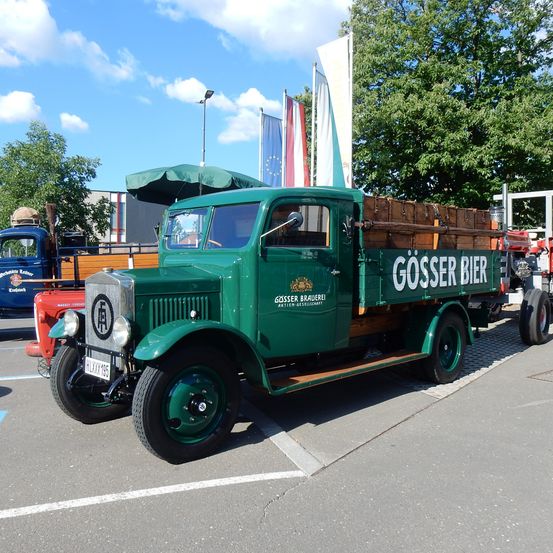 Ein alter grüner Lastwagen mit den Worten 'Gösser Bier' an der Seite steht auf einem Parkplatz. Der Lastwagen hat Holzkisten auf der Rückseite und steht neben einem roten Fahrzeug.