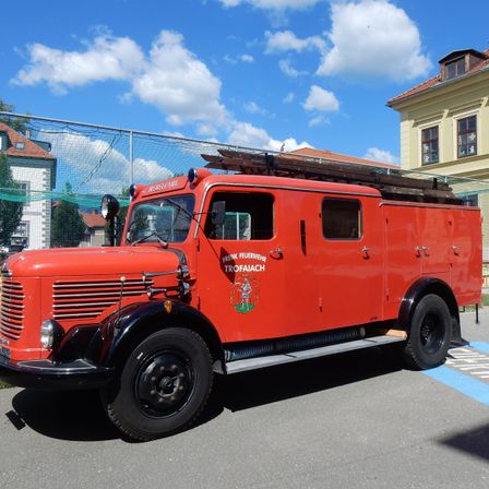 Ein roter Vintage-Feuerwehrwagen mit dem Wort 'Trofaiach' an der Seite, geparkt vor einem Gebäude mit einem Zaun und einem blauen Himmel im Hintergrund.