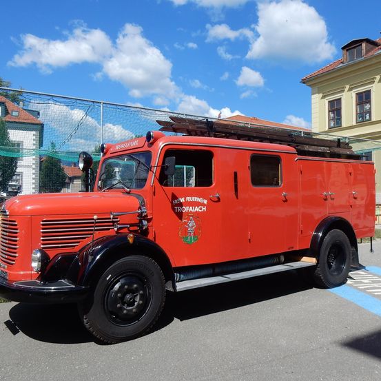 Ein roter Vintage-Feuerwehrwagen mit dem Wort 'Trofaiach' an der Seite, geparkt vor einem Gebäude mit einem Zaun und einem blauen Himmel im Hintergrund.