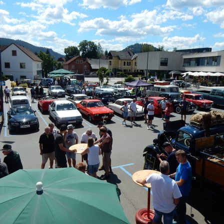Ein Parkplatz voller klassischer Autos, mit Menschen, die herumlaufen und einige stehen bei einem Tisch mit einem Regenschirm. Gebäude und Bäume im Hintergrund.
