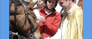 Ein Priester segnet ein Pferd, während eine Frau und ein Mann in roten Jacken zusehen. Die Frau trägt einen Helm.