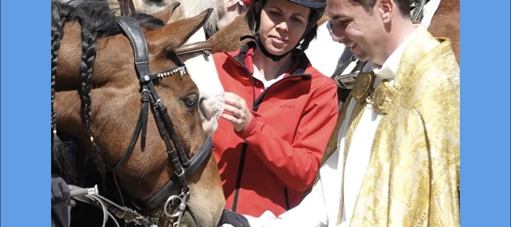 Ein Priester segnet ein Pferd, während eine Frau und ein Mann in roten Jacken zusehen. Die Frau trägt einen Helm.