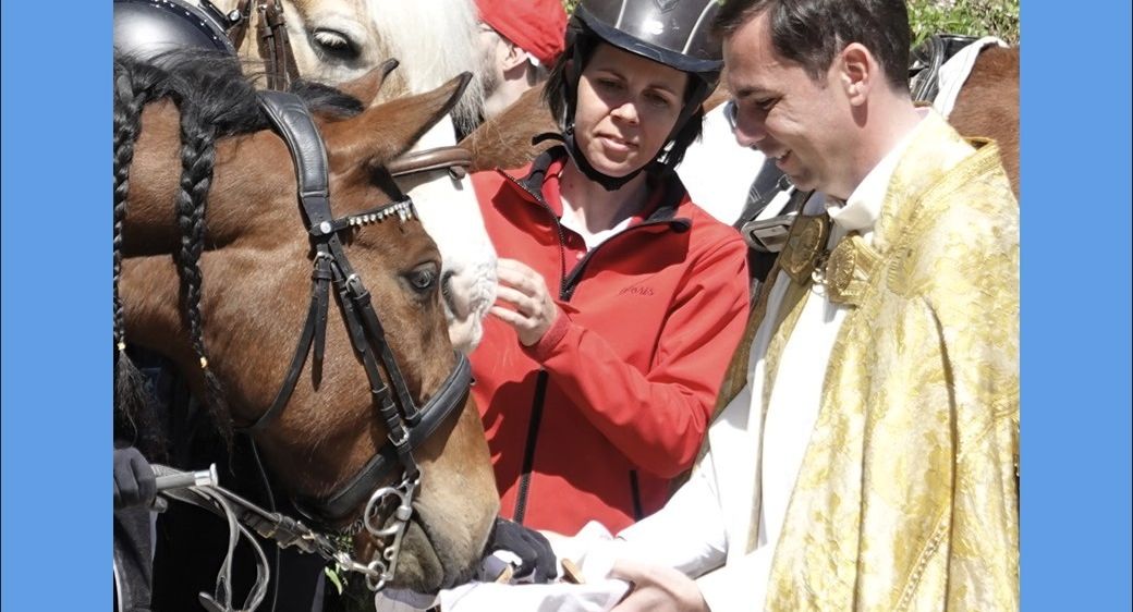 Ein Priester segnet ein Pferd, während eine Frau und ein Mann in roten Jacken zusehen. Die Frau trägt einen Helm.