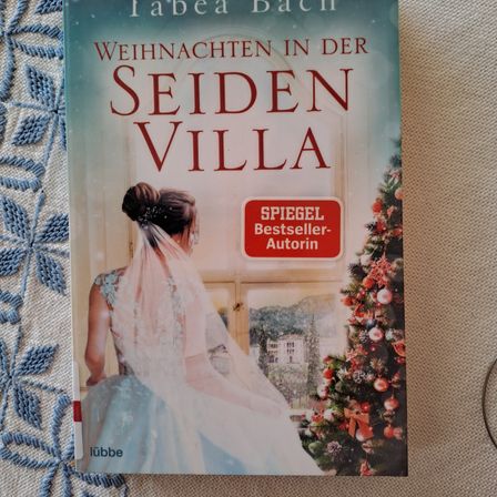 Ein Buch mit dem Titel 'Weihnachten in der Seiden Villa' zeigt eine Braut in einem Hochzeitskleid und einen Weihnachtsbaum auf dem Cover. Das Cover liegt auf einer gemusterten Oberfläche.