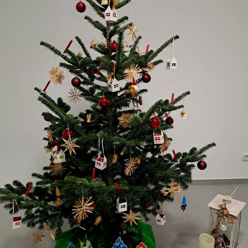 Ein geschmückter Weihnachtsbaum mit roten Kugeln, kleinen weißen Häusern und goldenen Sternen steht in einem Raum. Ein Laterne steht daneben.