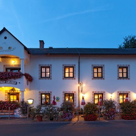 Die Vorderseite eines weißen Gebäudes mit dem Schild 'Hof Gruber' ist bei Nacht beleuchtet. Es hat einen Schornstein, mehrere Fenster und dekorative Muster. Davor stehen Blumen und Pflanzen sowie geschlossene Regenschirme.