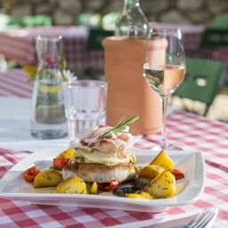 Ein Gericht mit Schweinekotelett mit Käse und Gemüse auf einem weißen Teller, serviert mit einem Glas Weißwein und einer Flasche Wasser auf einer rot-weiß karierten Tischdecke.