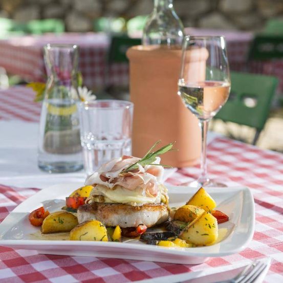 Ein Gericht mit Schweinekotelett mit Käse und Gemüse auf einem weißen Teller, serviert mit einem Glas Weißwein und einer Flasche Wasser auf einer rot-weiß karierten Tischdecke.
