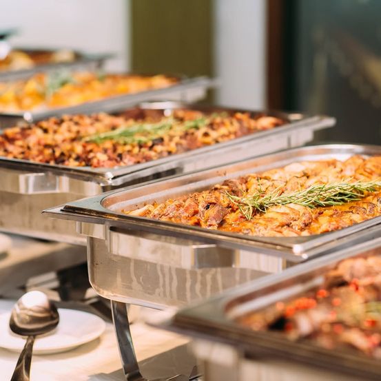 Eine Reihe von Edelstahl-Buffet-Tabletts mit verschiedenen Gerichten, mit frischen Kräutern garniert, auf einer weißen Oberfläche.