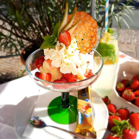 Eine Glasschale mit einem Erdbeerdessert, überzogen mit Schlagsahne, einer Scheibe Birne und einem Cracker. Daneben ein Glas Limonade und eine Schüssel mit Erdbeeren.