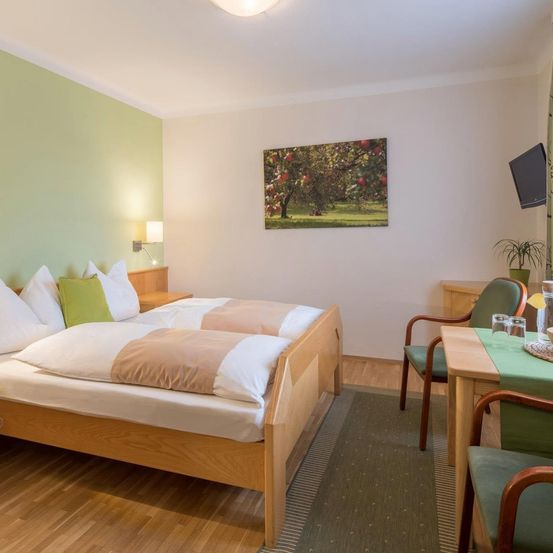 Ein gemütliches Schlafzimmer mit zwei Betten, einer grünen Wand und einem Gemälde von Äpfeln. Ein Fernseher ist an der Wand montiert, und ein Tisch mit einer Pflanze steht in der Ecke.