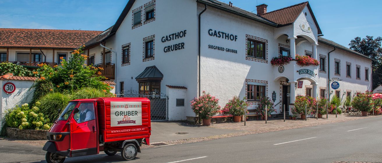 A red vehicle is driving down the road in front of Gasthof Gruber, a white building with red roof. The building has red flowers on the windows.