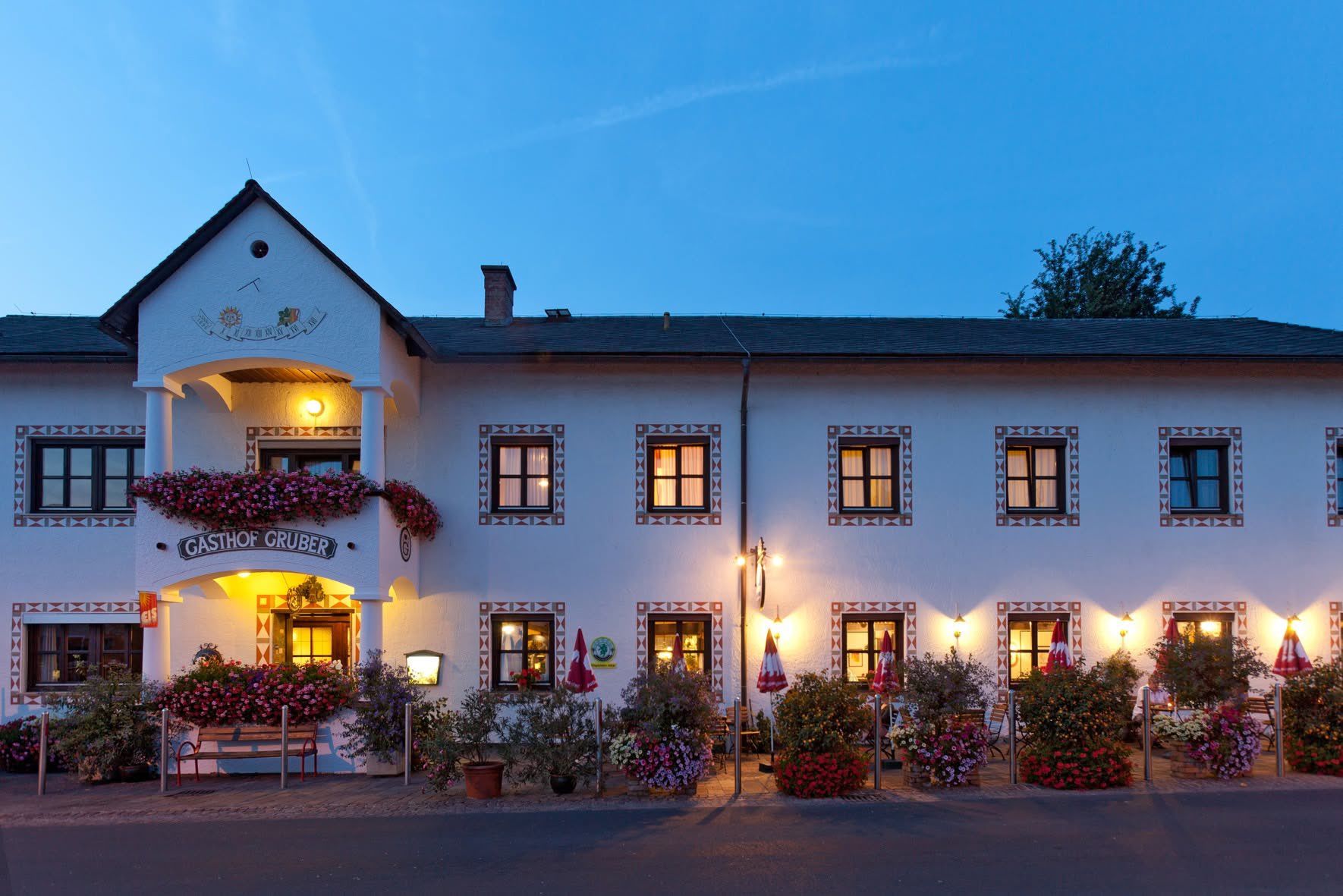 A white building with multiple windows and decorative trims is lit up at dusk. The building has a name board and flowers on the front. There are umbrellas and plants in front.