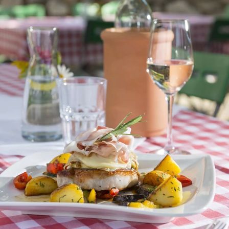 Ein Gericht mit Schweinefleisch, überzogen mit Käse, Schinken und Rosmarin, serviert mit Gemüse und Wein auf einer rot-weiß karierten Tischdecke.