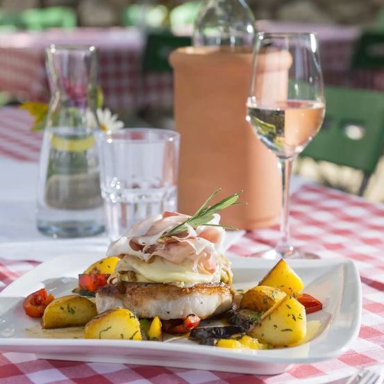 A dish of pork topped with cheese, ham, and rosemary, served with vegetables and wine on a red-and-white-checkered tablecloth.