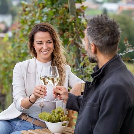 Eine lächelnde Frau und ein Mann, die Weingläser halten, sitzen eng beieinander in einem Weinberg. Sie scheinen einen Moment der Entspannung und Unterhaltung zu genießen.