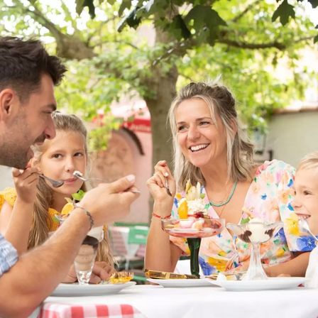 Eine Familie genießt gemeinsam eine Mahlzeit in einem Außenrestaurant, wobei der Mann und die beiden Mädchen lächelnd Dessert essen.