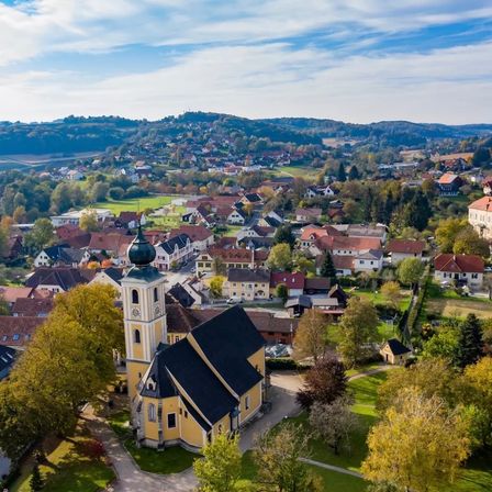 Luftaufnahme eines Dorfes mit vielen Häusern und einer Kirche. Die Kirche hat einen Turm und eine grüne Kuppel. Das Dorf ist von Bäumen und Gras umgeben.