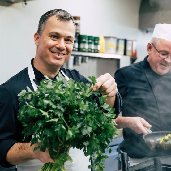 Two chefs in a kitchen. One holds a bunch of parsley, the other cooks with a frying pan.