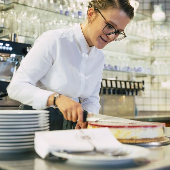 A woman in a white shirt and glasses cuts a cake in a restaurant with shelves and glasses in the background.
