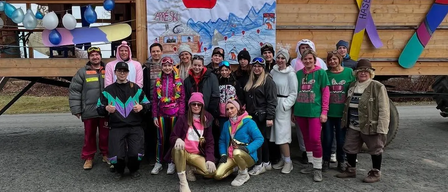 Eine Gruppe von Menschen in Winterkleidung posiert vor einer hölzernen Struktur mit einem Plakat an einem bewölkten Tag.