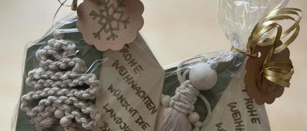 Eine Person hält zwei Pakete mit Dekorationsartikeln. Jedes Paket ist in durchsichtige Plastikfolie eingewickelt und mit goldenen Bändern gebunden. Die Artikel sind mit weißem Garn gestrickt und haben ein Schneeflocken-Etikett. Der Text auf dem Etikett lautet 'ROH WEIHNACHTEN WUNSCH DE LANDJUGEND BILDEIN'.