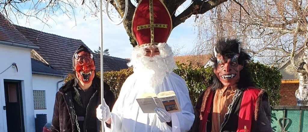 Drei Personen in Kostümen stehen vor einem Baum. Die zentrale Figur trägt eine weiße Robe, einen roten Bischofshut und hält ein Buch. Die Person auf der linken Seite trägt eine rote Maske und Handschuhe. Die Person auf der rechten Seite trägt ein rotes und schwarzes Outfit. Hinter ihnen steht ein Haus und ein grüner Zaun.