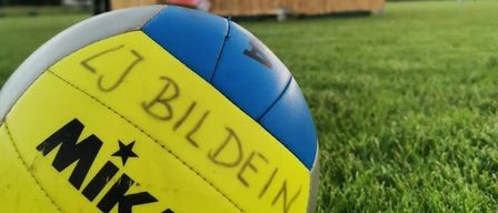 Ein Volleyball mit der Aufschrift 'Mikasa LJ Bilderin' liegt auf einem Rasenfeld. Im Hintergrund steht eine Hütte mit grünem Vordach hinter einem Volleyballnetz.