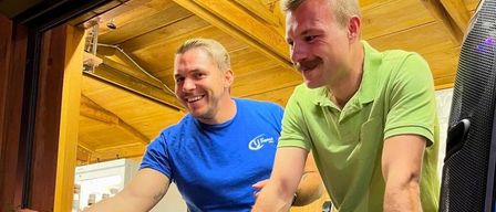 Zwei Männer lächeln und stehen in einem Raum mit Holzwänden. Der Mann auf der linken Seite trägt ein blaues T-Shirt und hat ein Tattoo auf dem Arm. Der Mann auf der rechten Seite trägt ein grünes Poloshirt und hat einen Schnurrbart.