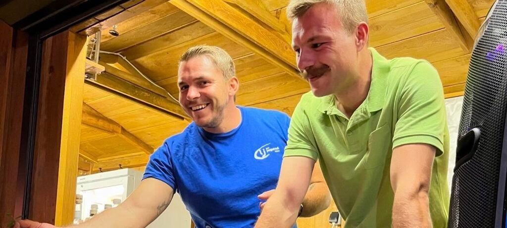 Zwei Männer lächeln und stehen in einem Raum mit Holzwänden. Der Mann auf der linken Seite trägt ein blaues T-Shirt und hat ein Tattoo auf dem Arm. Der Mann auf der rechten Seite trägt ein grünes Poloshirt und hat einen Schnurrbart.