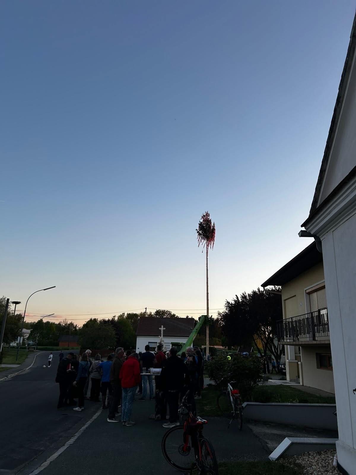 At dusk, a group of people are gathered on a street, with a tall pole adorned with red flowers, near a house with a balcony, and a bicycle parked nearby.