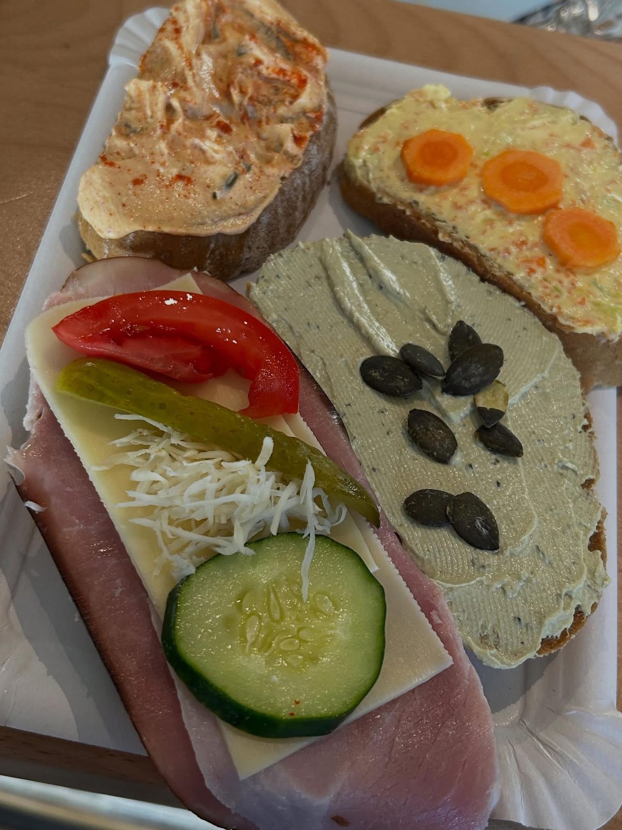 Ein Teller mit vier verschiedenen Sandwiches. Eines hat Schinken, Käse, Tomate und Gurke. Ein anderes hat eine Aufstrich mit Kürbiskernen. Das dritte hat einen Aufstrich mit Karotten. Das letzte hat einen Aufstrich mit Kräutern.