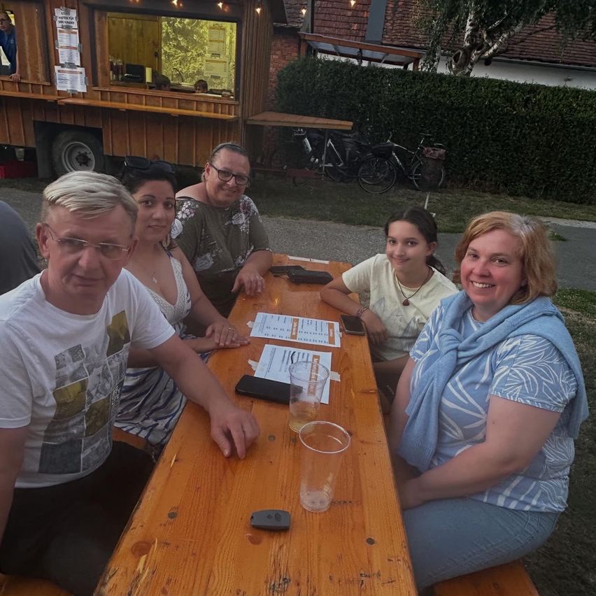 Eine Gruppe von Menschen sitzt um einen Holztisch und posiert lächelnd für ein Foto. Hinter ihnen steht ein Imbisswagen und Fahrräder.