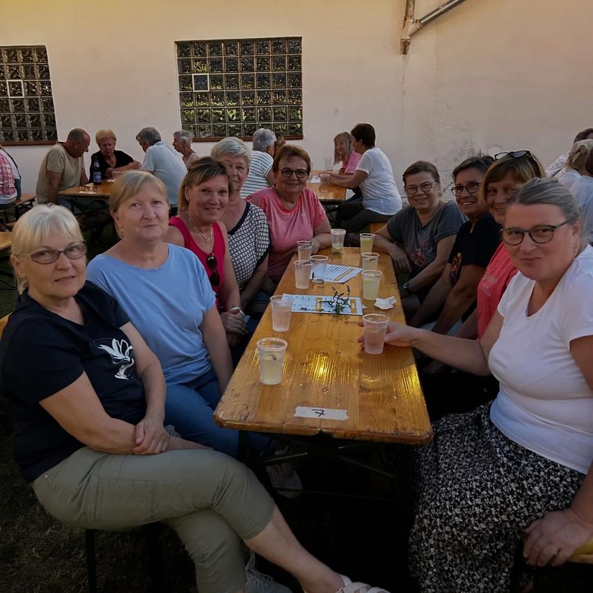 Eine Gruppe von Frauen und Männern sitzt um einen Tisch herum, lächelt und posiert für ein Foto. Sie befinden sich in einem Außenbereich mit einer weißen Wand und Glasfenstern.